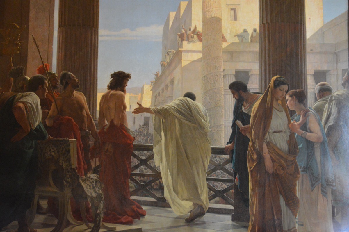 A painting: on a palace balcony Pilate, in a pale robe, gestures toward the scourged Jesus, crowned with thorns, before a crowd below